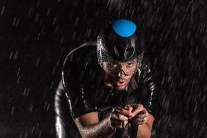 A triathlete braving the rain as he cycles through the night, preparing himself for the upcoming marathon. The blurred raindrops in the foreground and the dark, moody atmosphere in the background add photo