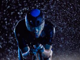 A triathlete braving the rain as he cycles through the night, preparing himself for the upcoming marathon. The blurred raindrops in the foreground and the dark, moody atmosphere in the background add photo