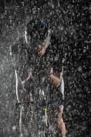 A triathlete braving the rain as he cycles through the night, preparing himself for the upcoming marathon. The blurred raindrops in the foreground and the dark, moody atmosphere in the background add photo