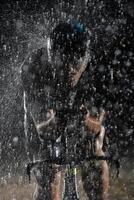 A triathlete braving the rain as he cycles through the night, preparing himself for the upcoming marathon. The blurred raindrops in the foreground and the dark, moody atmosphere in the background add photo