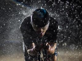 A triathlete braving the rain as he cycles through the night, preparing himself for the upcoming marathon. The blurred raindrops in the foreground and the dark, moody atmosphere in the background add photo