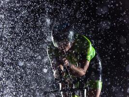 A triathlete braving the rain as he cycles through the night, preparing himself for the upcoming marathon. The blurred raindrops in the foreground and the dark, moody atmosphere in the background add photo