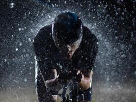 A triathlete braving the rain as he cycles through the night, preparing himself for the upcoming marathon. The blurred raindrops in the foreground and the dark, moody atmosphere in the background add photo