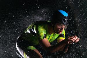A triathlete braving the rain as he cycles through the night, preparing himself for the upcoming marathon. The blurred raindrops in the foreground and the dark, moody atmosphere in the background add photo