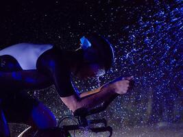 A triathlete braving the rain as he cycles through the night, preparing himself for the upcoming marathon. The blurred raindrops in the foreground and the dark, moody atmosphere in the background add photo
