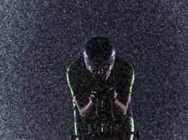A triathlete braving the rain as he cycles through the night, preparing himself for the upcoming marathon. The blurred raindrops in the foreground and the dark, moody atmosphere in the background add photo