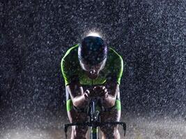 A triathlete braving the rain as he cycles through the night, preparing himself for the upcoming marathon. The blurred raindrops in the foreground and the dark, moody atmosphere in the background add photo