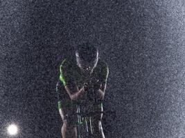 A triathlete braving the rain as he cycles through the night, preparing himself for the upcoming marathon. The blurred raindrops in the foreground and the dark, moody atmosphere in the background add photo