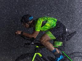 A triathlete braving the rain as he cycles through the night, preparing himself for the upcoming marathon. The blurred raindrops in the foreground and the dark, moody atmosphere in the background add photo