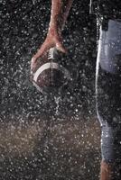 Close up of American Football Athlete Warrior Standing on a Field focus on his Helmet and Ready to Play. Player Preparing to Run, Attack and Score Touchdown. Rainy Night with Dramatic lens flare photo