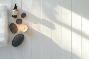 A minimalist composition featuring massage stones, essential oil bottles, and a lit candle arranged on a white wooden table. photo