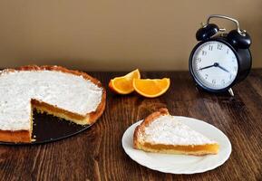 Piece of homemade pie with lemon and orange filling, covered with powdered sugar on a wooden table near orange fruit and alarm clock photo