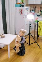 Little girl with a big teddy bear stands near the table in the light of a lamp on a tripod photo