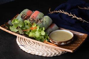 Fresh spring rolls filled with vibrant vegetables and garnished with sesame seeds are served on wooden plate with side of dipping sauce, creating healthy and appetizing dish photo
