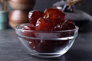 glass bowl filled with glossy, dark red dates sits on dark surface, showcasing their rich texture and inviting appearance. background features blurred kitchen items, adding cozy atmosphere photo