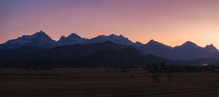 Silhouette of Bavarian Alps at dusk with Neuschwanstein Castle faintly illuminated. Open fields, scattered trees, and gradient sky complete the scene. photo