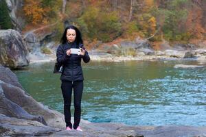A young woman takes a photo near a rocky mountain river with a rapid flow