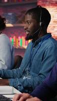 Vertical Portrait of smiling technical support specialist in call center office doing troubleshooting. Upbeat african american IT worker fixing customer connectivity issue, using headphones, camera B video