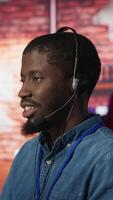 Vertical Portrait of smiling technical support specialist in call center office doing troubleshooting. Upbeat african american IT worker fixing customer connectivity issue, using headphones, camera A video
