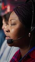 Vertical Call center employee talks in headphones mic, handling customer inquiries and complaints in office. African american customer service representative assisting customers with issues, camera B close up video