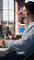 Vertical Happy call center agent in office helping customers by answering questions, solving issues. Smiling woman in customer support workspace in front of PC using headset mic to talk with clients, camera B video