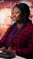Vertical Upbeat call center agent provides guidance to customers, answering questions. Jolly African american customer center worker at PC desk walking clients through technical solutions during call, camera B video