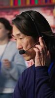 Vertical Call center senior agent providing step by step guidance to help customers. Asian experienced customer center worker at PC desk walking clients through technical solutions during call, camera A video