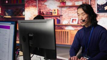 Call center coworkers dealing with upset customers, speaking with them online using headset at office desk. Asian man using PC to do customer support job, handling clients complaints, camera B video