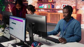 Smiling call center agent dealing with unhappy customers, speaking with them using audio headset at office desk. Customer support worker handling clients complaints in company workspace, camera B video