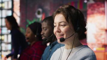 Happy call center agent in office helping customers by answering questions, solving issues. Smiling woman in customer support workspace in front of PC using headset mic to talk with clients, camera A video