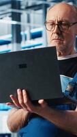 Vertical Elderly engineer uses laptop in smart factory to test automated systems, ensuring reliability. Experienced technician uses notebook to identify bottlenecks and optimize equipment, camera A video