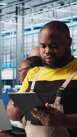 Vertical Smart factory engineer oversees operation of robotic arms and assembly lines on facility floor. Industry 4.0 plant african american man monitors system performance and troubleshoots issues, camera A video