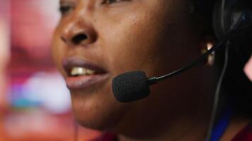 Closeup of call center employee talks in headphones mic, handling customer inquiries and complaints in office. African american customer service representative assists customers with issues, camera A video