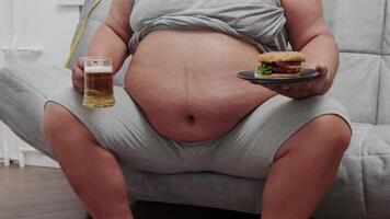 un de edad mediana hombre tiene un bocadillo en el sofá. un grasa hombre se sienta en un sofá y tiene un cerveza y come un hamburguesa video
