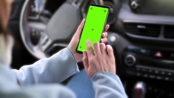 a woman holding a smartphone with a green screen on the background of the steering wheel in the car. video