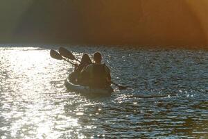 A young couple enjoying an idyllic kayak ride in the middle of a beautiful river surrounded by forest greenery in sunset time photo
