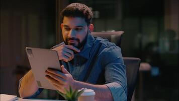 Smiling Indian Arabian man satisfied businessman ethnic male guy business entrepreneur in dark office late night working using digital tablet gadget addict browsing device online chatting internet app video