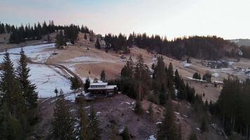 Mountain cabin surrounded by snow-dusted trees, serene setting with rolling hills and scattered houses in the background, peaceful winter dusk atmosphere video