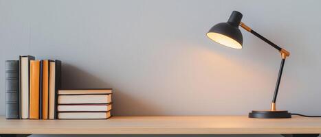 Modern desk lamp with a black and gold design illuminating a wooden table with neatly arranged books against a gray wall. A minimalistic and cozy reading or study space concept. photo