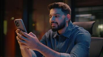 Indian ethnic man business employer in dark evening office late working typing mobile phone scrolling gadget addict night insomnia smiling male businessman guy browsing smartphone online order device video