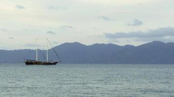A wide shot depicts a sailing ship powered by its engine, moving smoothly across the still, slightly rippled sea with its sails down. The vessel leaves a faint trail in the water under a soft, cloudy video