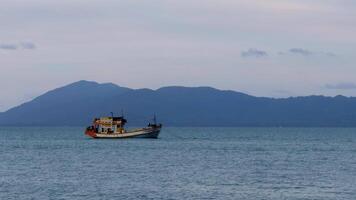 A colorful traditional fishing boat drifts on tranquil waters with a misty mountain range in the background. The soft lighting and pastel sky create a peaceful maritime scene video