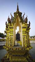 Closeup shot of an ornate gold Thai shrine with intricate carvings and architectural details. A white cat sits inside in the foreground contrasting against the gold decor with the temple in the video