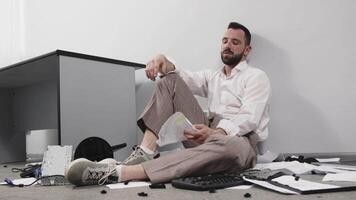 A businessman in a trashed office sits on the floor. Stress, chaos and business man with headache due to paperwork, reports. Concept of burnout, multitasking and emotional crisis video