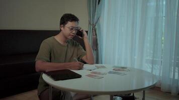 Financial worries a person wearing glasses sits at a table, penning notes while engaging in a phone call, with bills and a tablet scattered about, signifying financial difficulties. video