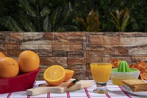 Glass of orange juice surrounded by orange juicer, red basket with oranges and plate with cookies against a brick wall background with green plants photo