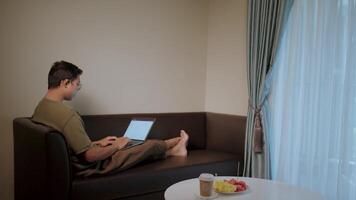 Man working from home on Sofa.A man relaxes on a comfortable couch, engrossed in his laptop, working from home with natural light streaming through the window. The scene exudes a sense of comfort. video