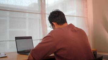 Focused on the task a man is intently working on his laptop, seen from behind. The natural light streaming in from the window illuminates his focus. video