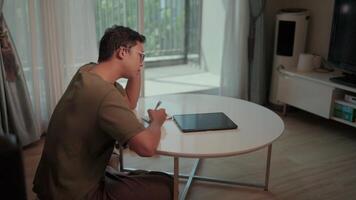 konzentriert Mann Arbeiten von heim. ein Mann sitzt beim ein klein runden Tabelle im seine Leben Zimmer, Konzentration intensiv wie er funktioniert auf ein Tablette und Notizblock. video