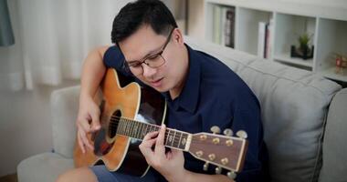 Handheld high angle view shot, Asian man sitting on sofa enjoying playing music with guitar and looking at music chords on laptop in living room with relaxation. video